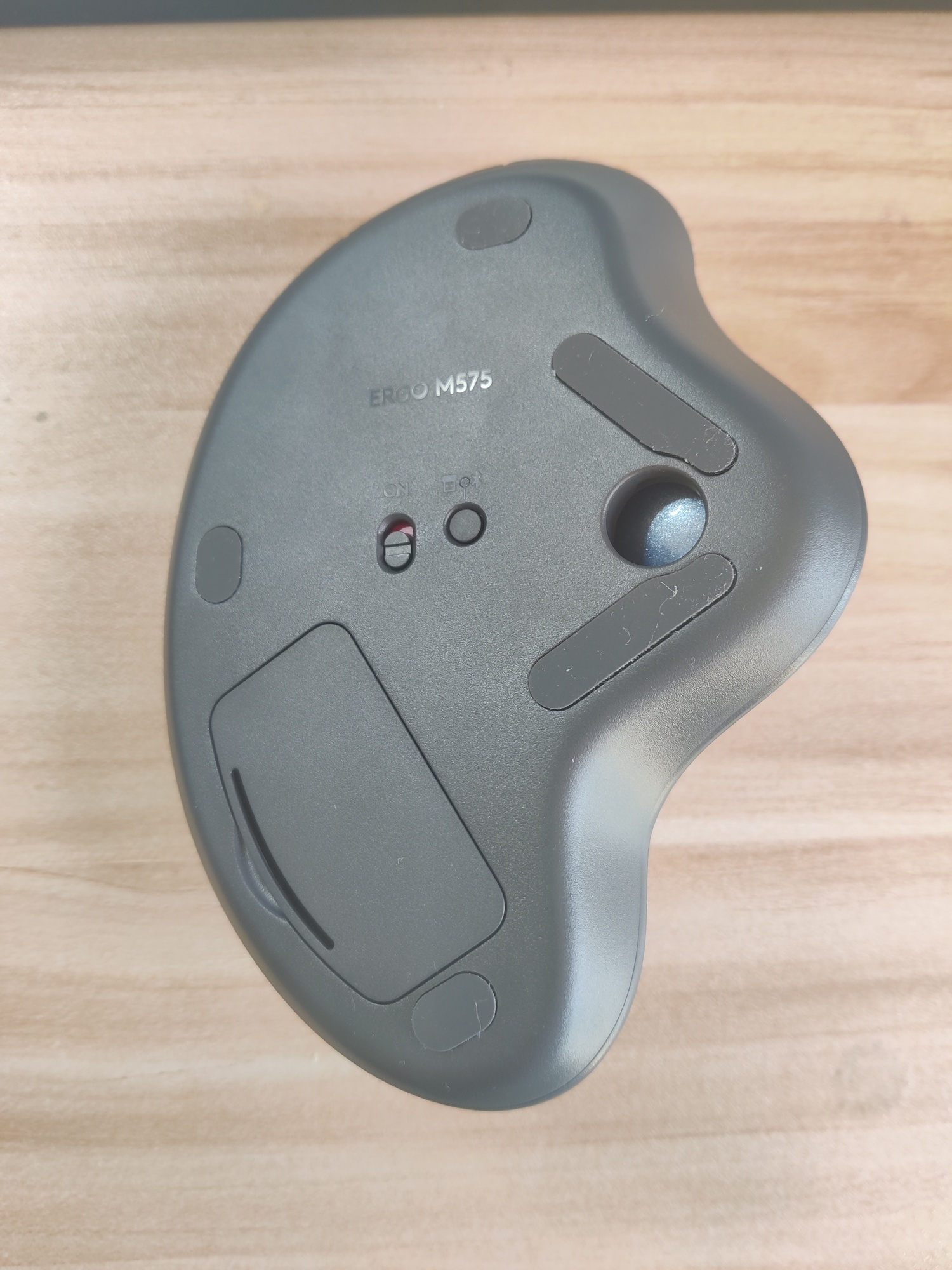Bottom view. A mostly flat surface except for an engraved "ERGO M575",
a few rubber pads, an on/off toggle switch, a dongle/bluetooth switch
button, a hole exposing bottom of trackball, and the lid of the battery
holder.