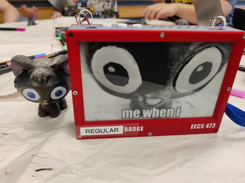 Figurine of a black cat next to an epaper screen displaying a goofy
image of him, captioned "me when i"